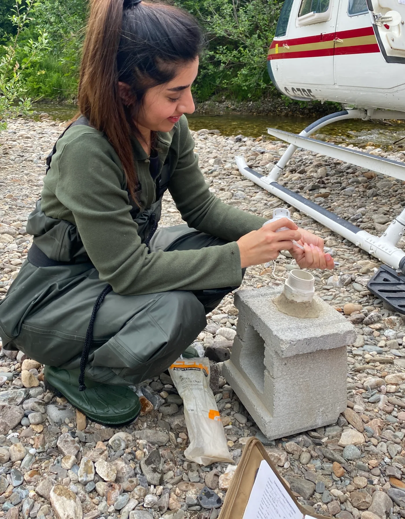 Behnoosh working with field equipment on a gravel bank beside a helicopter